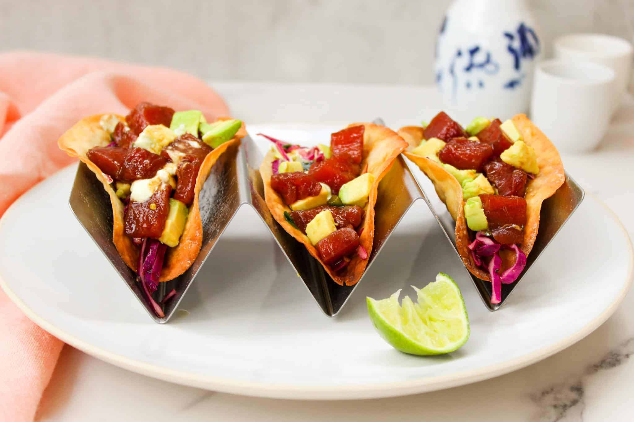 Poke tacos in taco stand on a white plate with lime wedge and sake.
