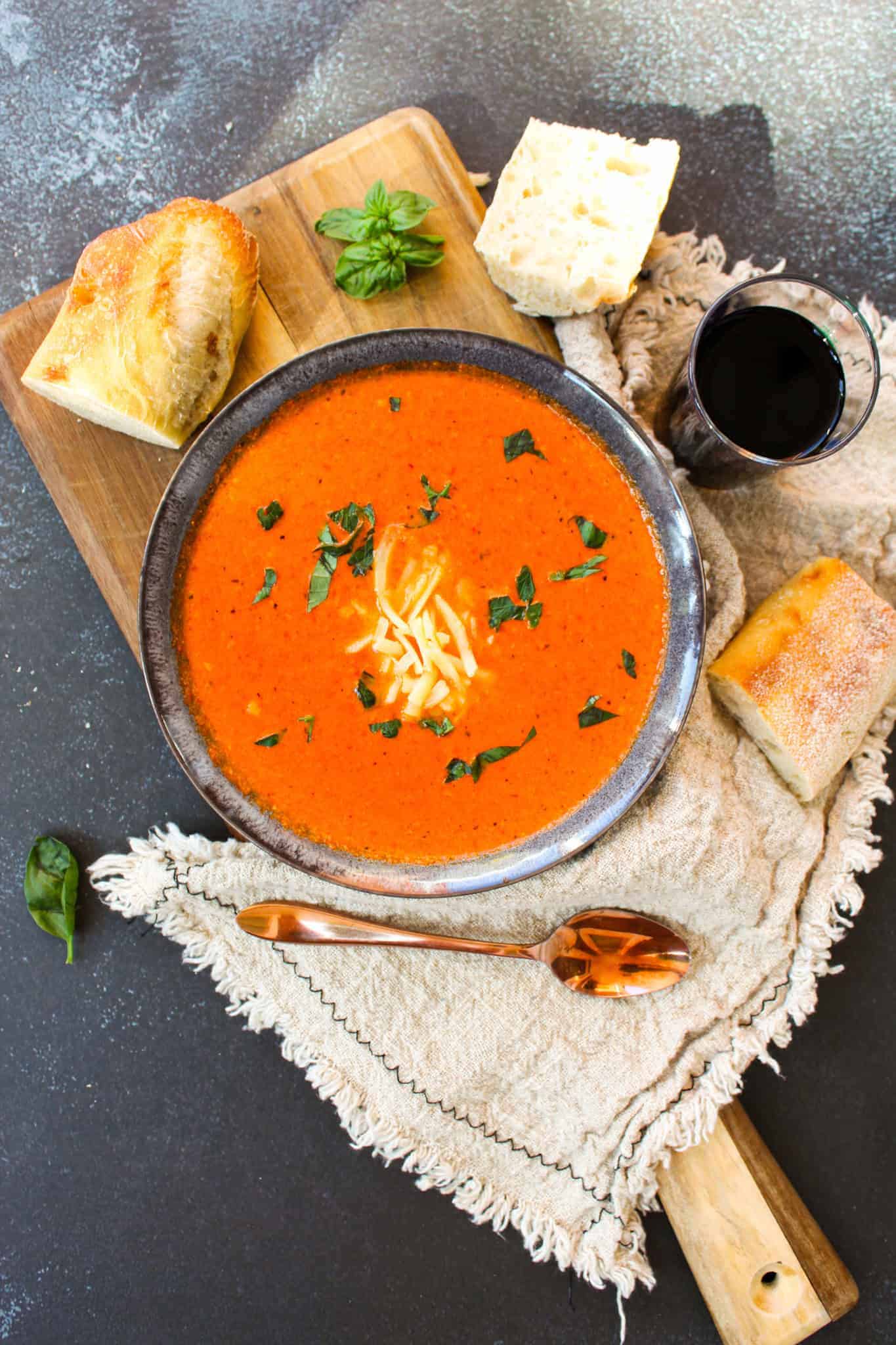 Roasted Red Pepper Gouda Soup in a bowl on a board next to bread, basil, and wine.