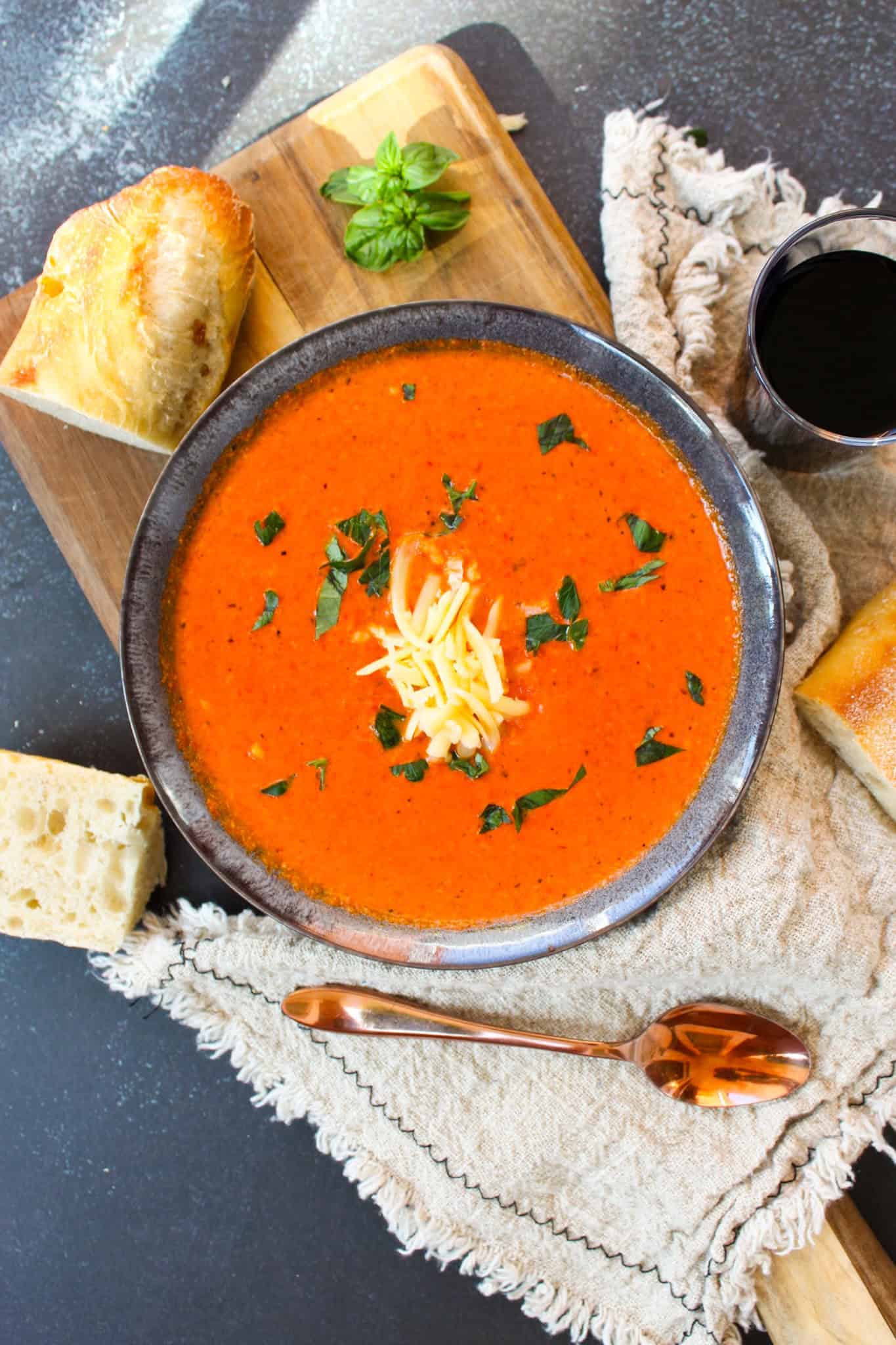 Rustic red soup topped with cheese next to bread, a spoon, and glass of wine.