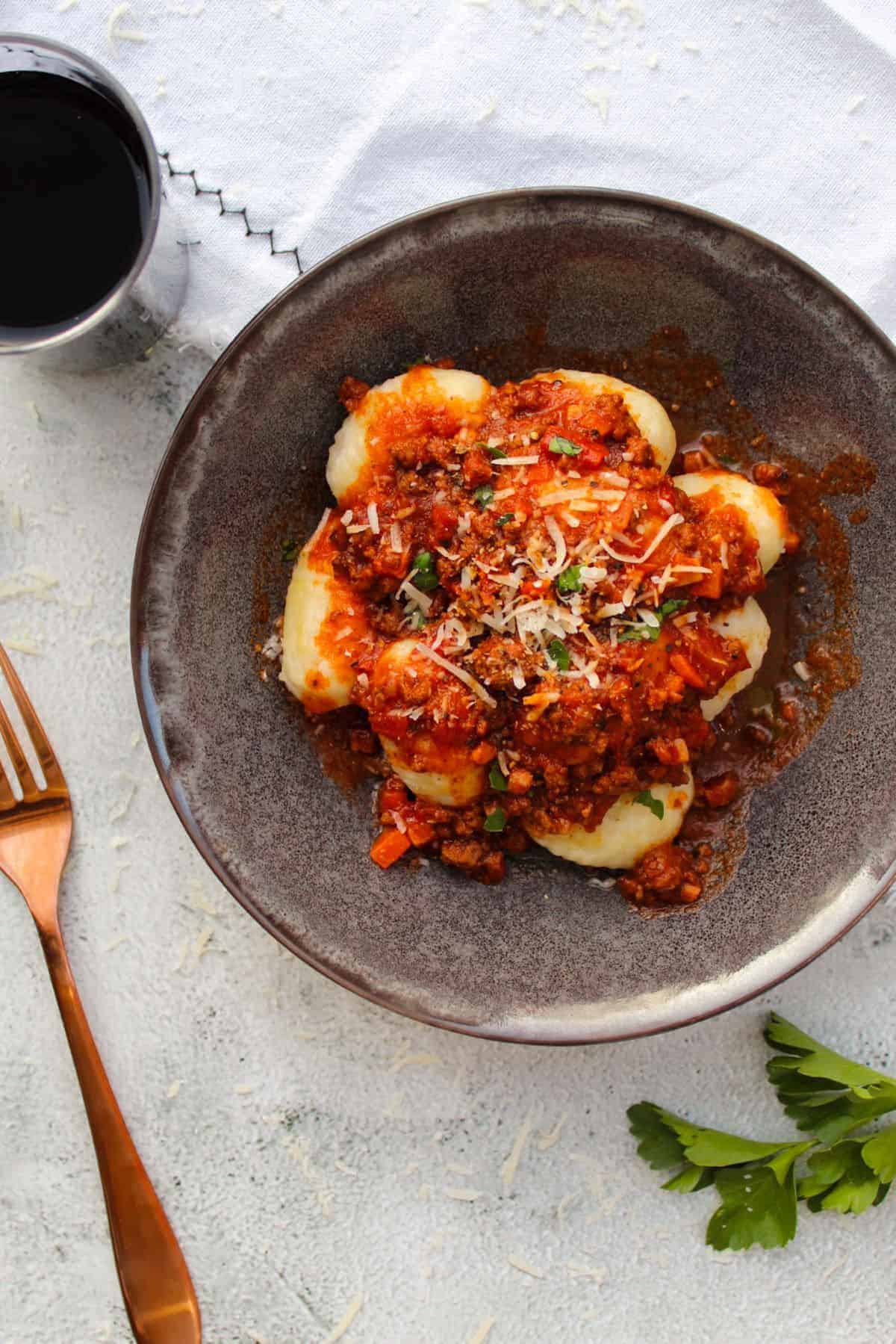 Gnocchi Bolognese in a charcoal bowl next to glass of red wine and fork.