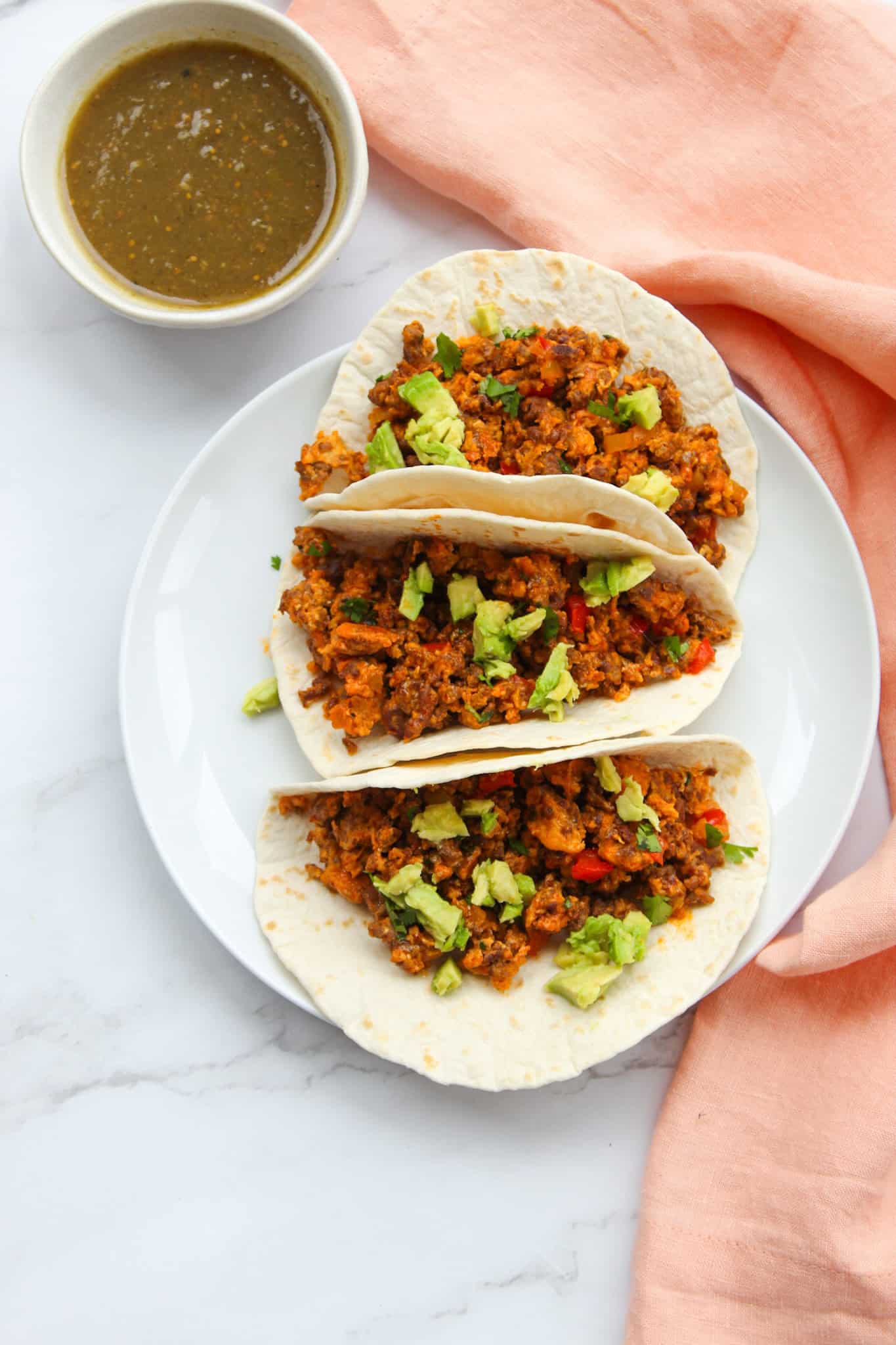 Breakfast tacos topped with avocado on a counter next to a bowl of salsa verde. 