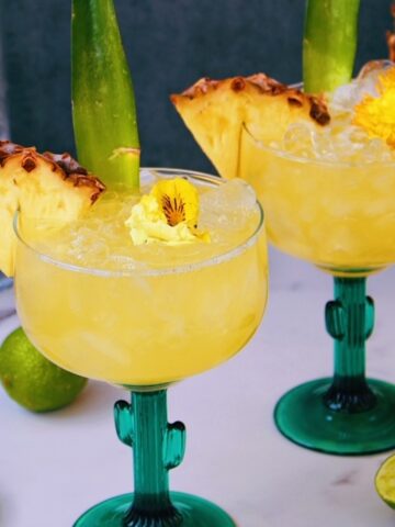 Two Pineapple Tequila Cocktails in cactus shaped glasses garnished with pineapple.