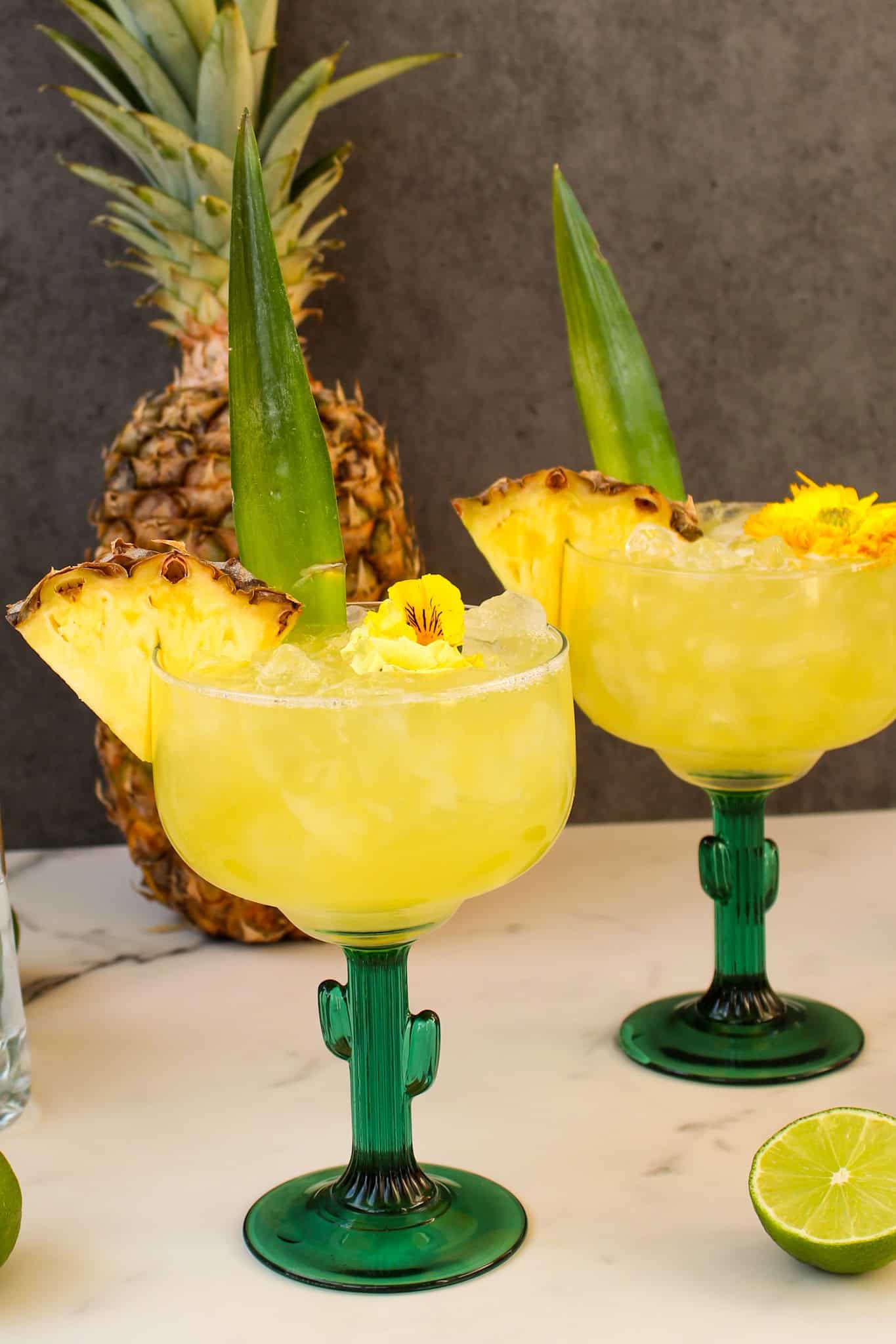Cactus shaped glasses with Pineapple Tequila cocktails inside next to lime and pineapple.