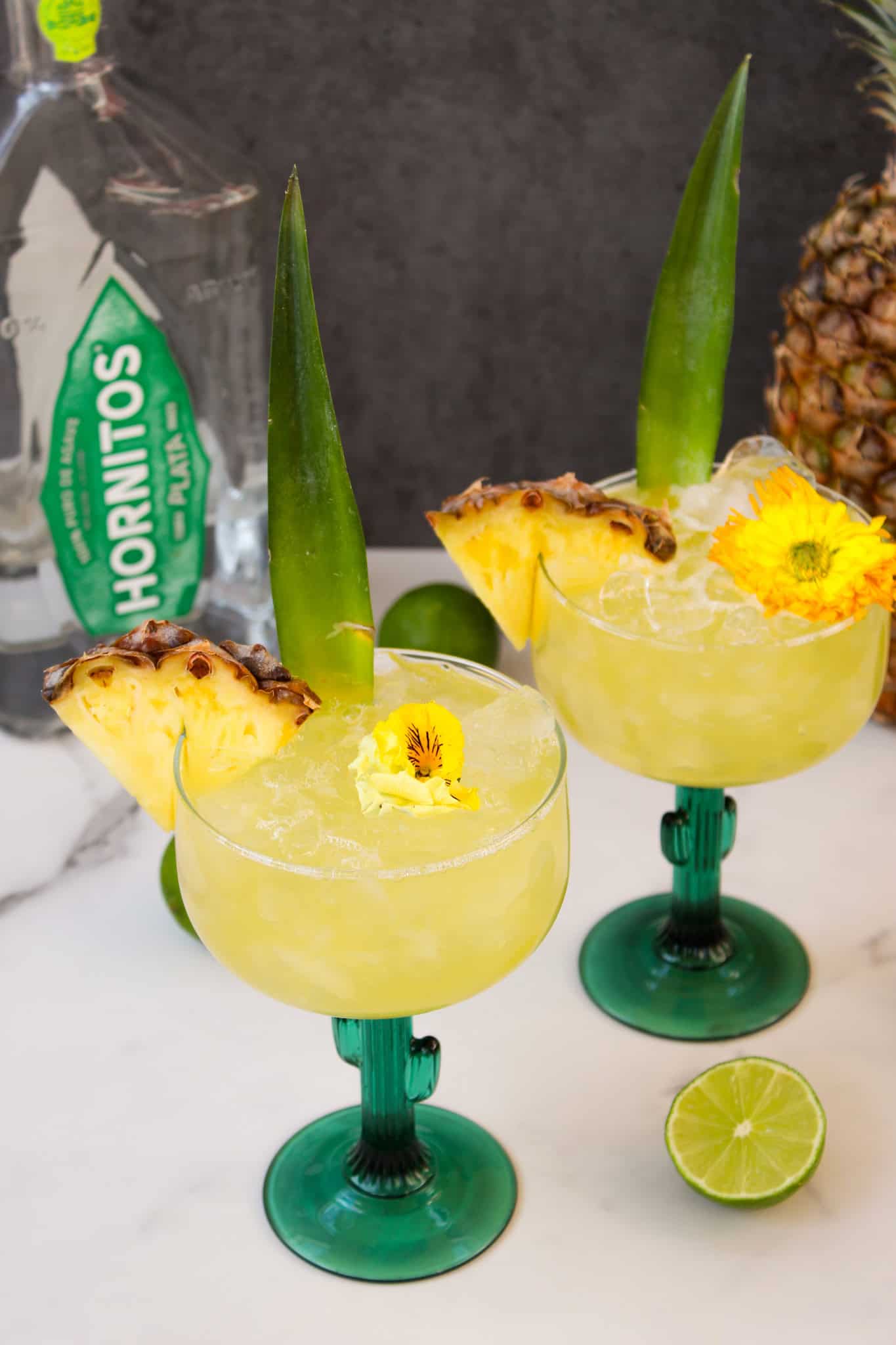 Tequila bottle next to two yellow margaritas and a whole pineapple.