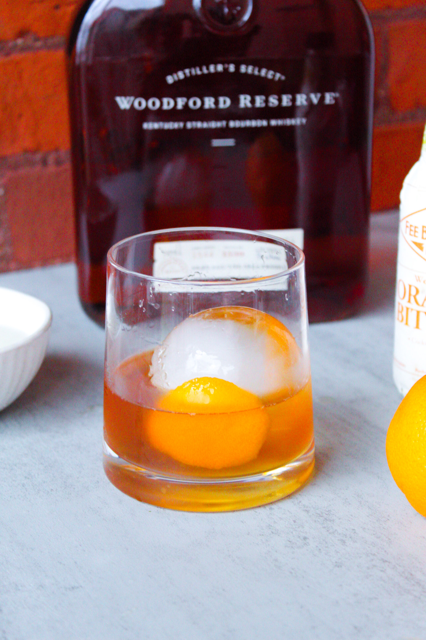 Old fashioned cocktail in rocks glass with ice ball sitting in front of bourbon.