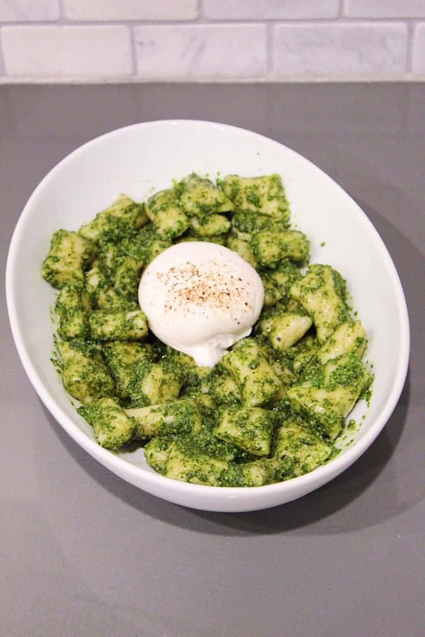 Homemade creamy basil pesto gnocchi in a large white serving bowl topped with burrata.