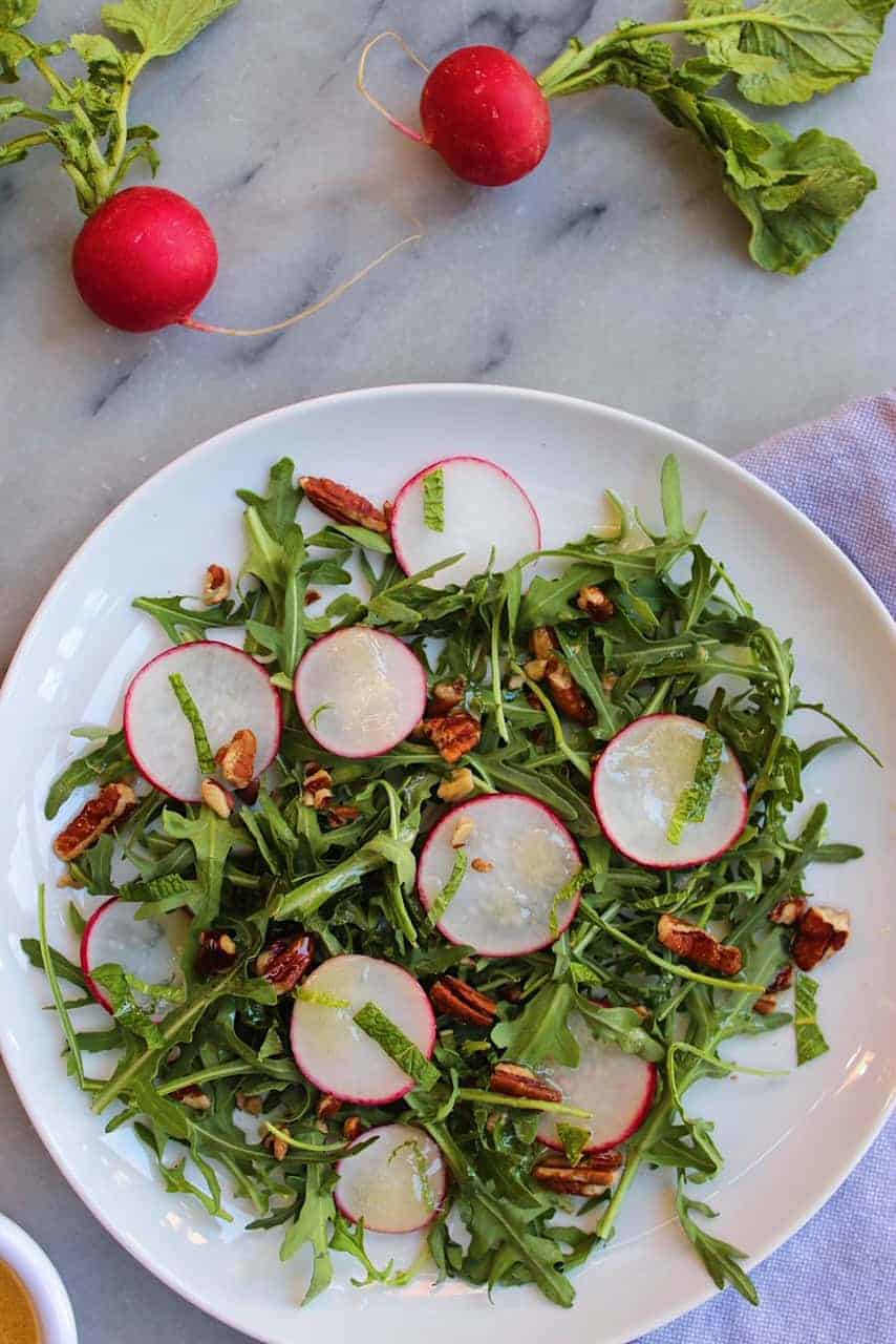 Radishes, pecans, mint, and arugula on a white plate over a marble countertop.