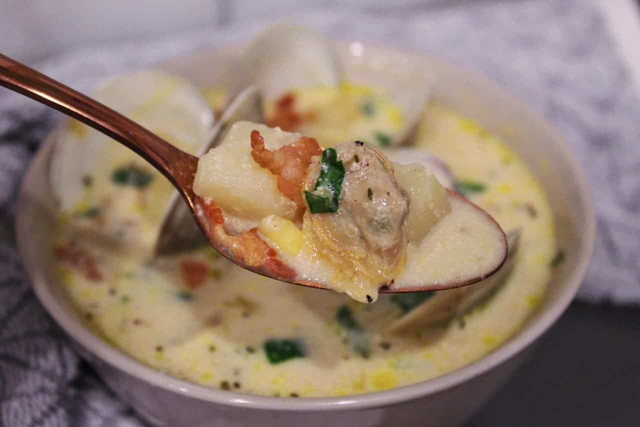 Close up spoonful of homemade New England clam chowder on a rose gold spoon with fresh clams, potato, bacon, and parsley.