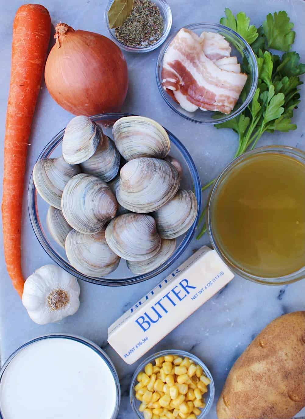 Overhead picture showing homemade clam chowder ingredients including carrot, onion, bacon, parsley, broth, potato, corn, heavy cream, butter, garlic, and fresh clams.