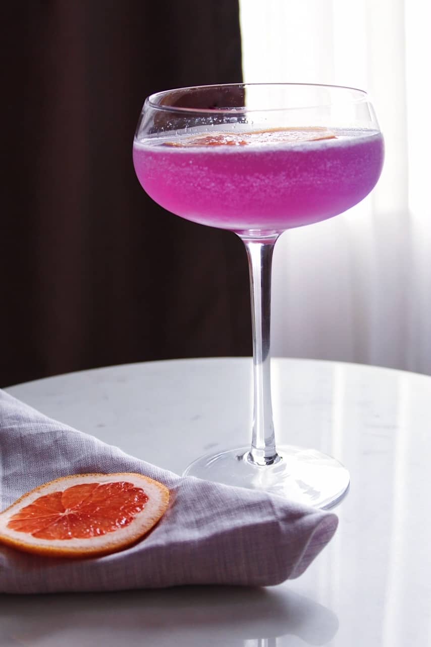 Pinkish purple Grapefruit Empress Gin cocktail in couple glass on marble table next to dish towel.