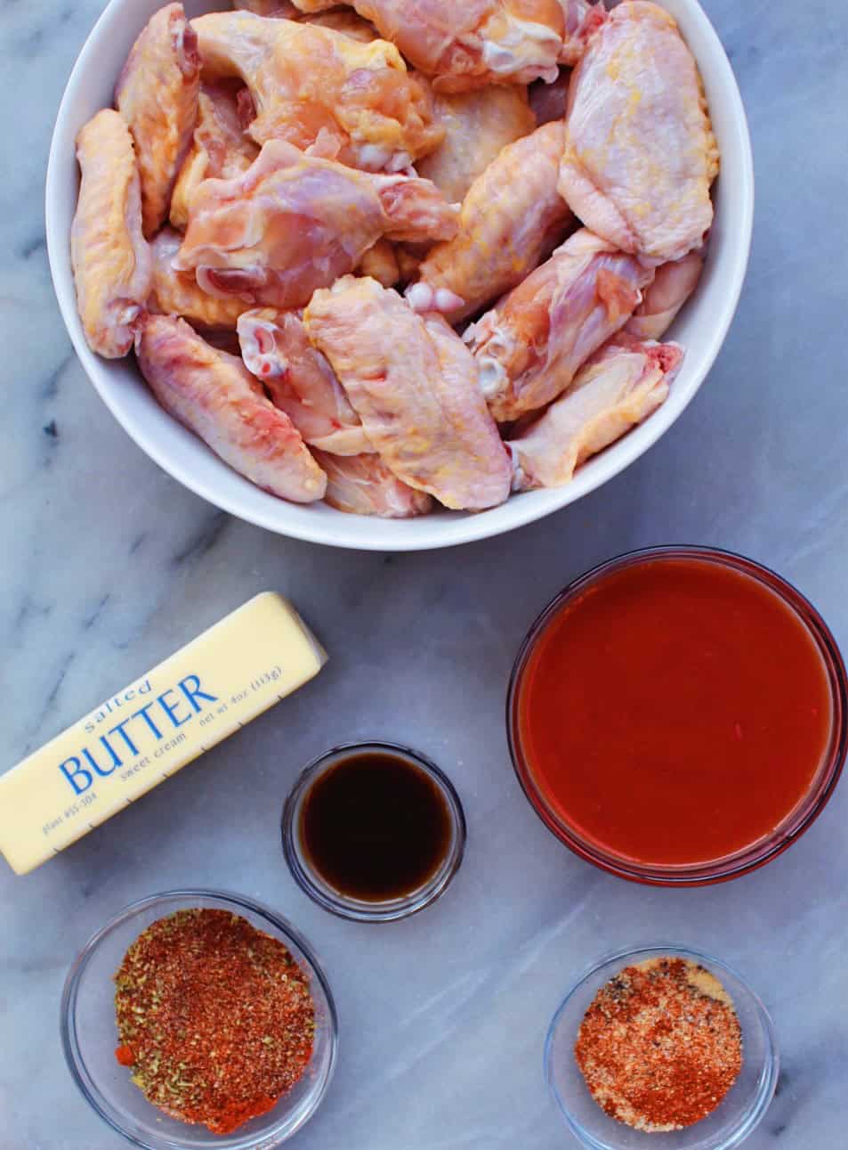 Separate bowls containing raw party wings, Frank's Red Hot sauce, Worcestershire sauce, spicy seasoning, and butter on a marble counter.