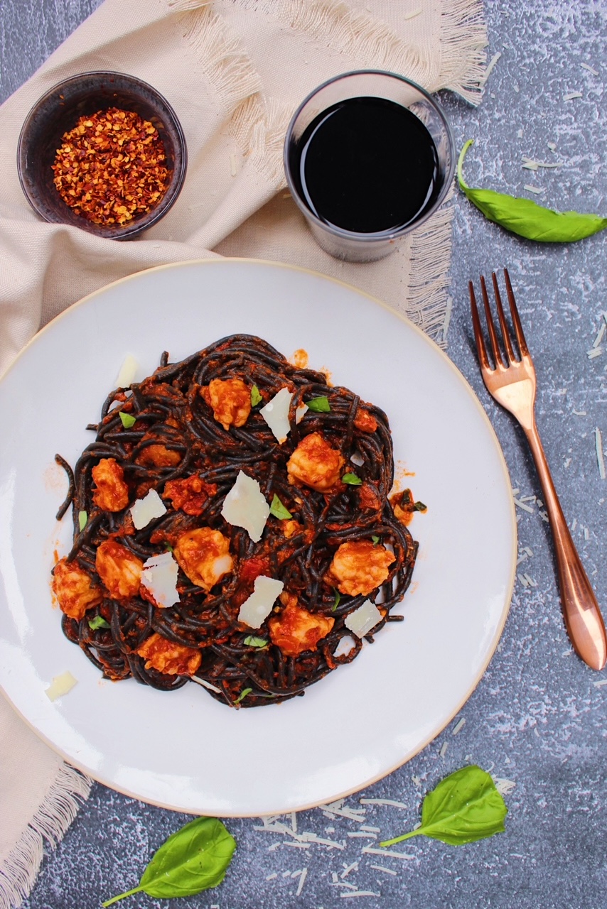 Overhead picture of squid ink pasta on a white plate with a glass of red wine and red pepper flakes on the side.