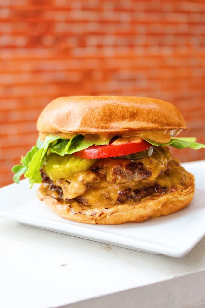 Burger on bun with toppings on a white plate in front of a brick wall.