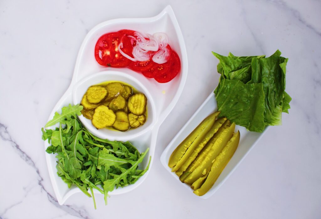 White tray holding arugula, pickles, tomato and onion next to a second white tray holding pickle spears and lettuce.