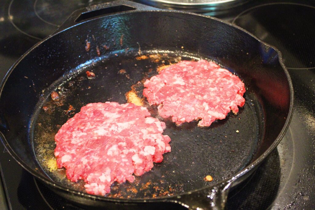 Two smash burgers cooking in a cast iron skillet.