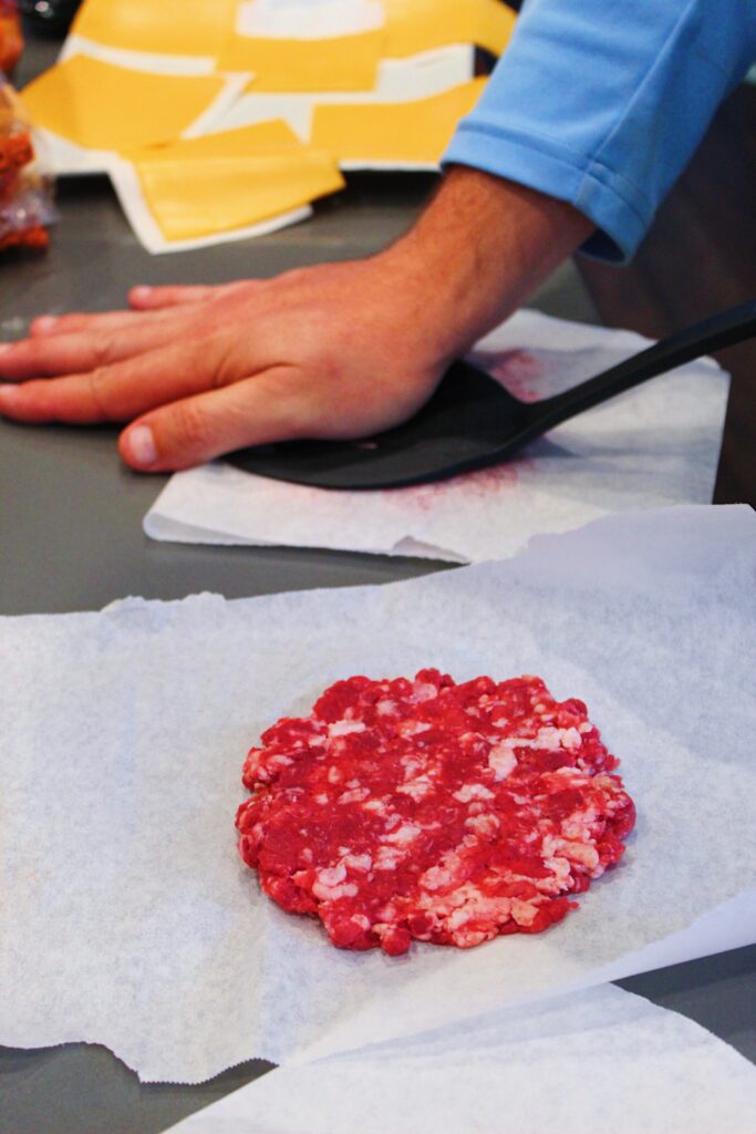 A hand pushing on a spatula over parchment paper to flatten burger patty with a flatted patty next to it.