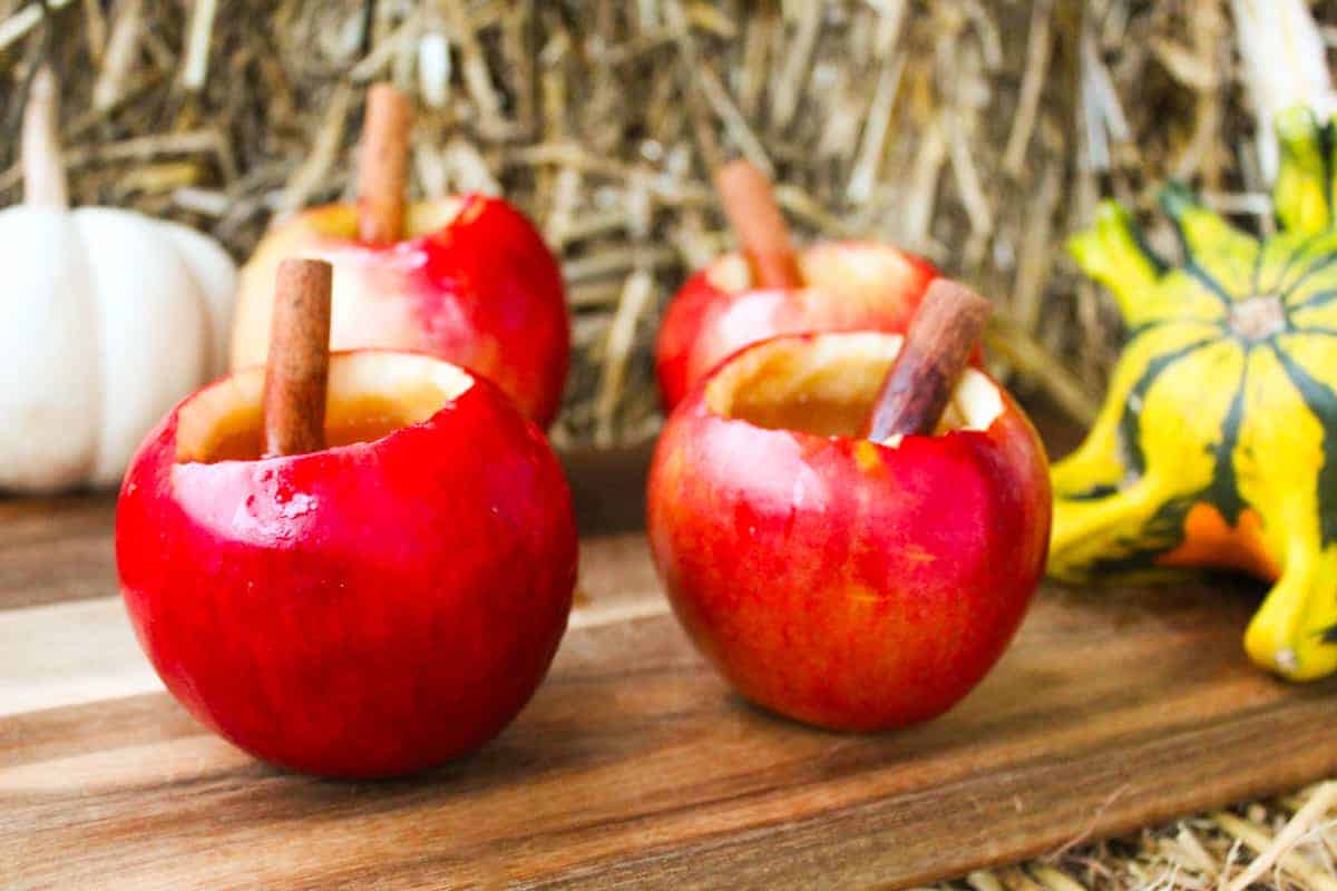 Apples containing spiked apple cider and cinnamon sticks on a wood surface next to pumpkins.