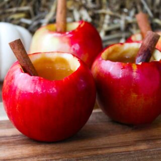 Four hollowed out apples filled with apple cider cocktail and cinnamon sticks on wood surface.