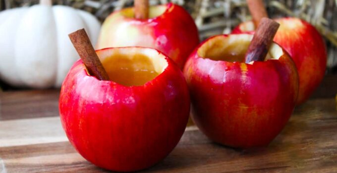 Four hollowed out apples filled with apple cider cocktail and cinnamon sticks on wood surface.