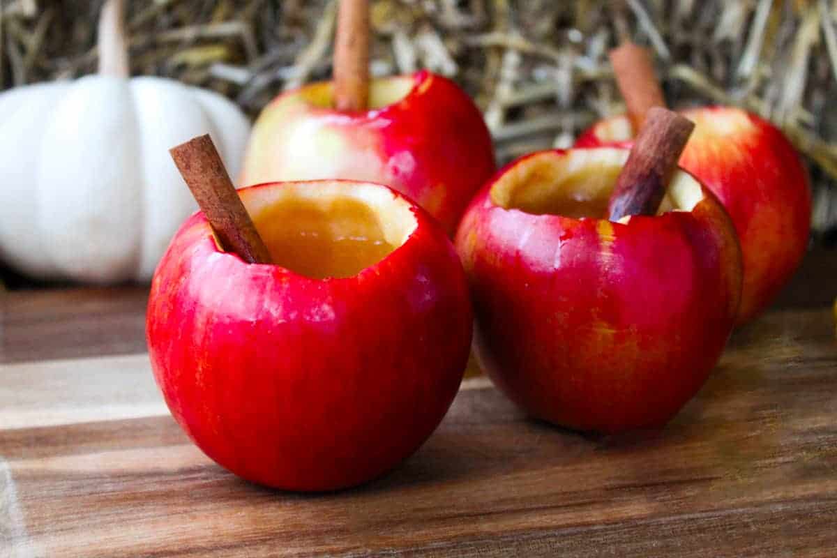 Four hollowed out apples filled with apple cider cocktail and cinnamon sticks on wood surface.
