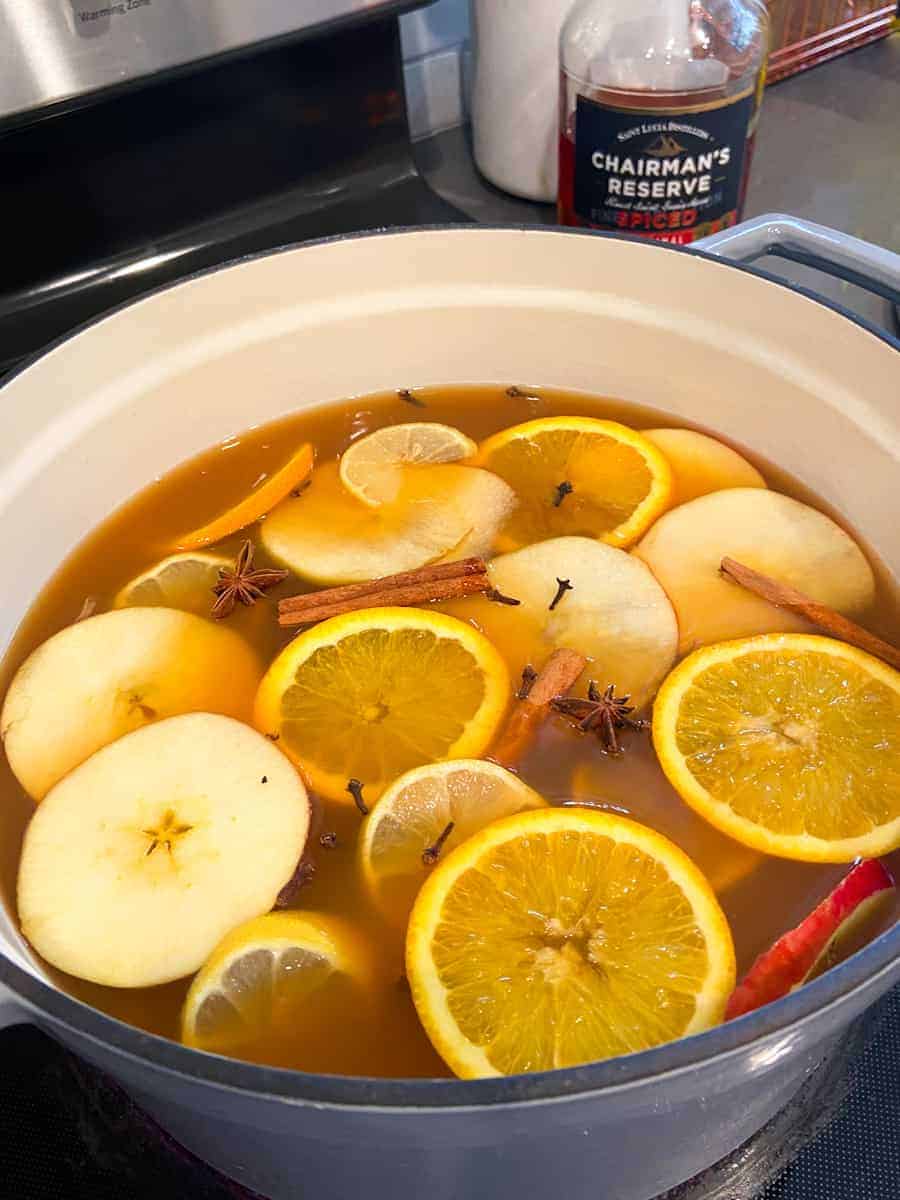 Apple cider simmering with spices, apple, orange, and lemon slices in large dutch oven next to bottle of spiced rum.